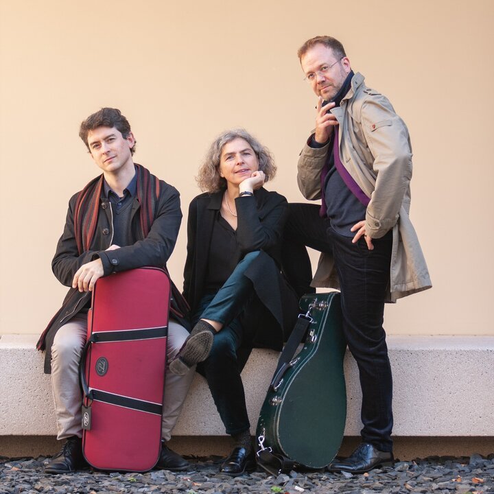 Drei Personen posieren selbstbewusst mit Musikinstrumentenkoffern vor einer schlichten beigen Wand.